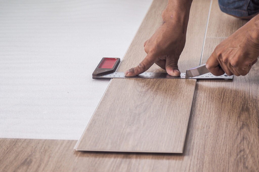 Person measuring and cutting a peel-and-stick flooring plank with a straight edge.