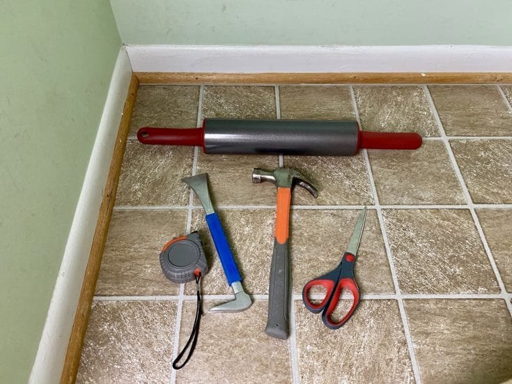 Basic tools for installing peel-and-stick floor tiles, including a roller, hammer, scraper, tape measure, and scissors.