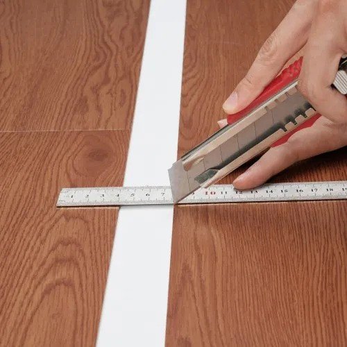 Person measuring and cutting a peel-and-stick flooring plank with a utility knife.